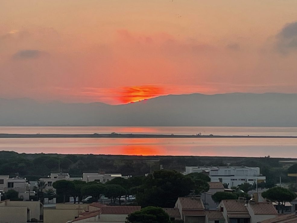 A sun set across the sea and towards the hills in the distance. The sky and sun are red, as is some of the sea.